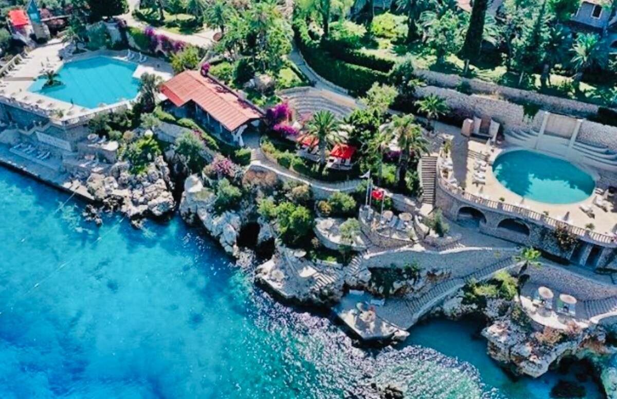 An aerial view displays a stunning coastal area featuring a large swimming pool alongside a hot tub, surrounded by vibrant greenery and colorful bougainvilleas. Sun loungers are positioned on the rocky shore, providing direct access to the crystal-clear waters of the Mediterranean.