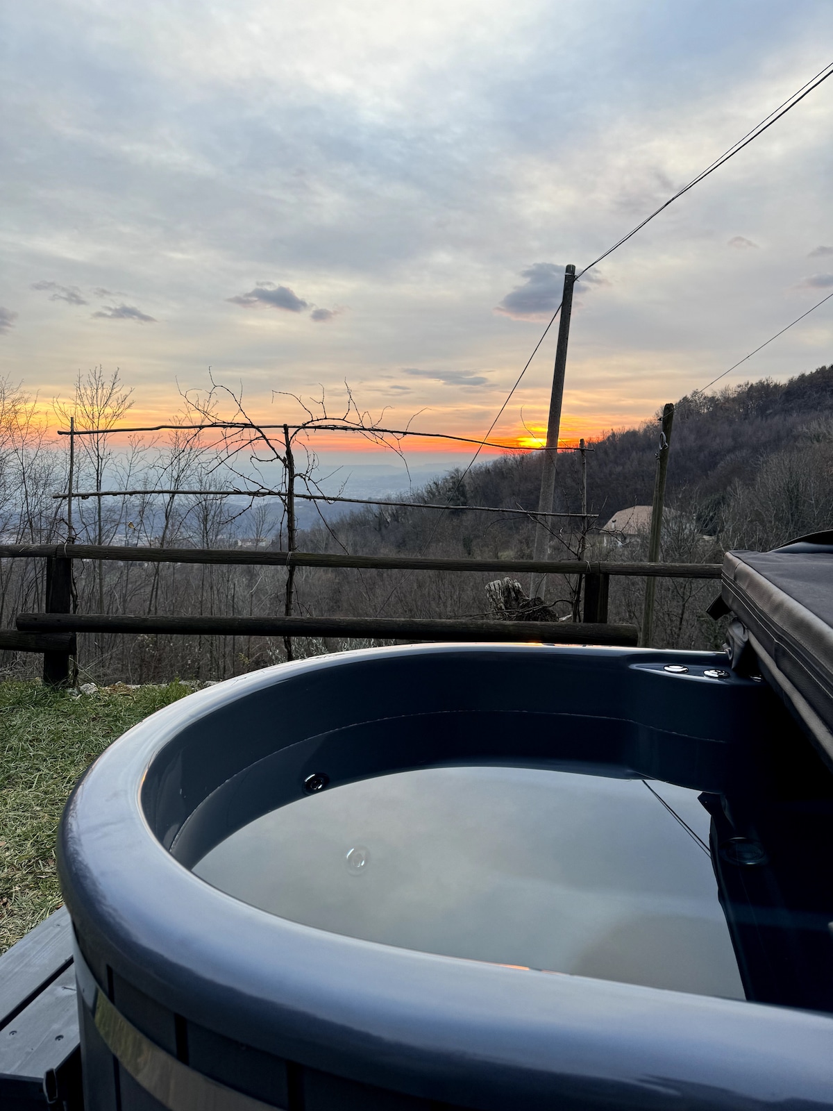 A hot tub is positioned outdoors, alongside a grassy area with a view of hills and trees. The sunset casts warm colors across the sky, enhancing the serene atmosphere. Wooden posts and power lines are visible in the background.