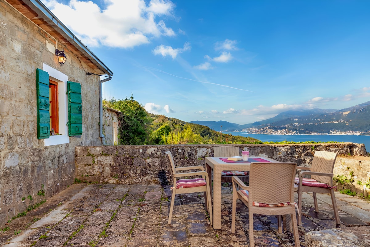 An outdoor terrace is shown, featuring a rectangular table surrounded by four chairs. The stone wall serves as a backdrop, with green shutters framing a nearby window. Scenic views of the mountains and sea are visible in the distance, under a partly cloudy sky.