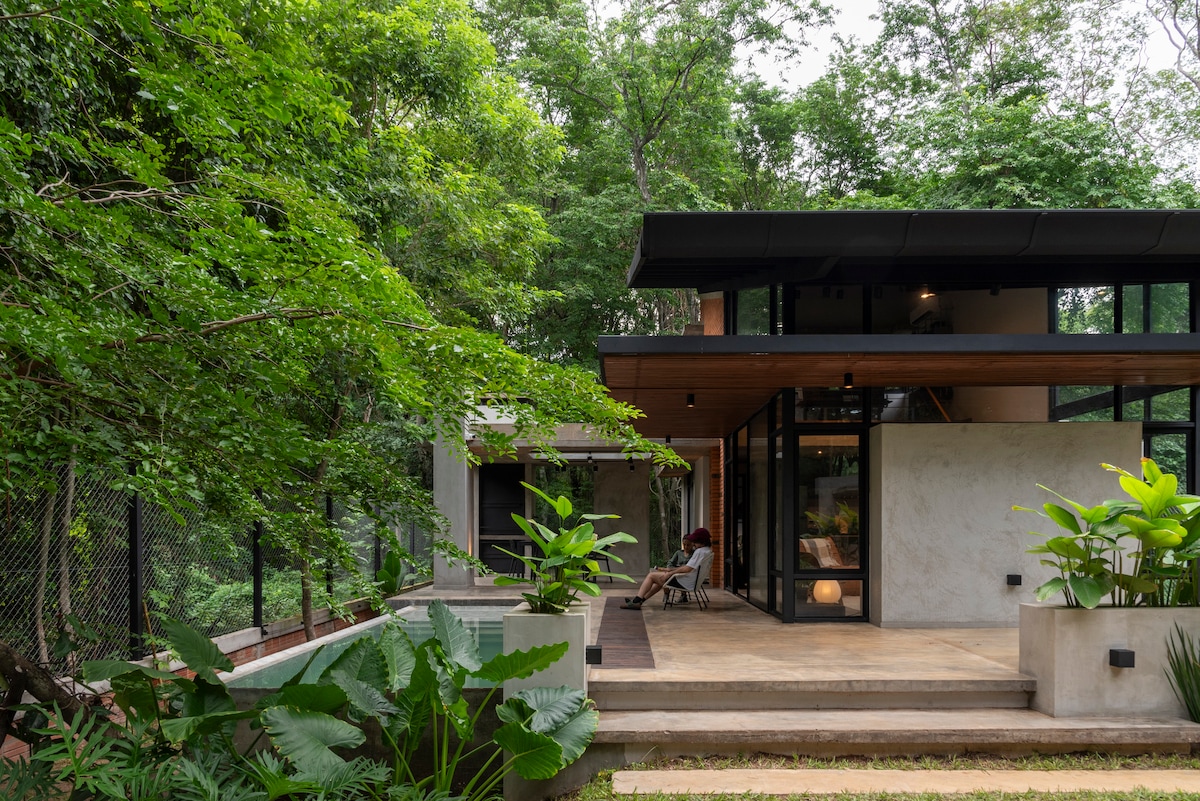 The exterior of the house reveals a modern design with large glass windows and a wooden overhang. Lush greenery surrounds the home, with scattered plants and trees creating a natural setting. A spacious outdoor area features a concrete patio, enhancing the connection with the forest.