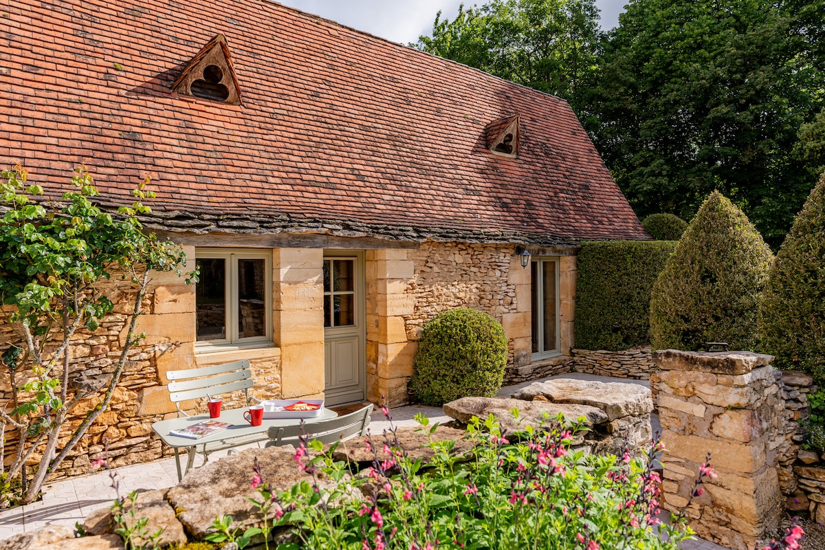 A charming stone building features a sloped roof with decorative elements. A small patio area is furnished with a table and chairs, surrounded by lush greenery and colorful flowers. The entrance is highlighted by double doors, providing access to the inviting interior.