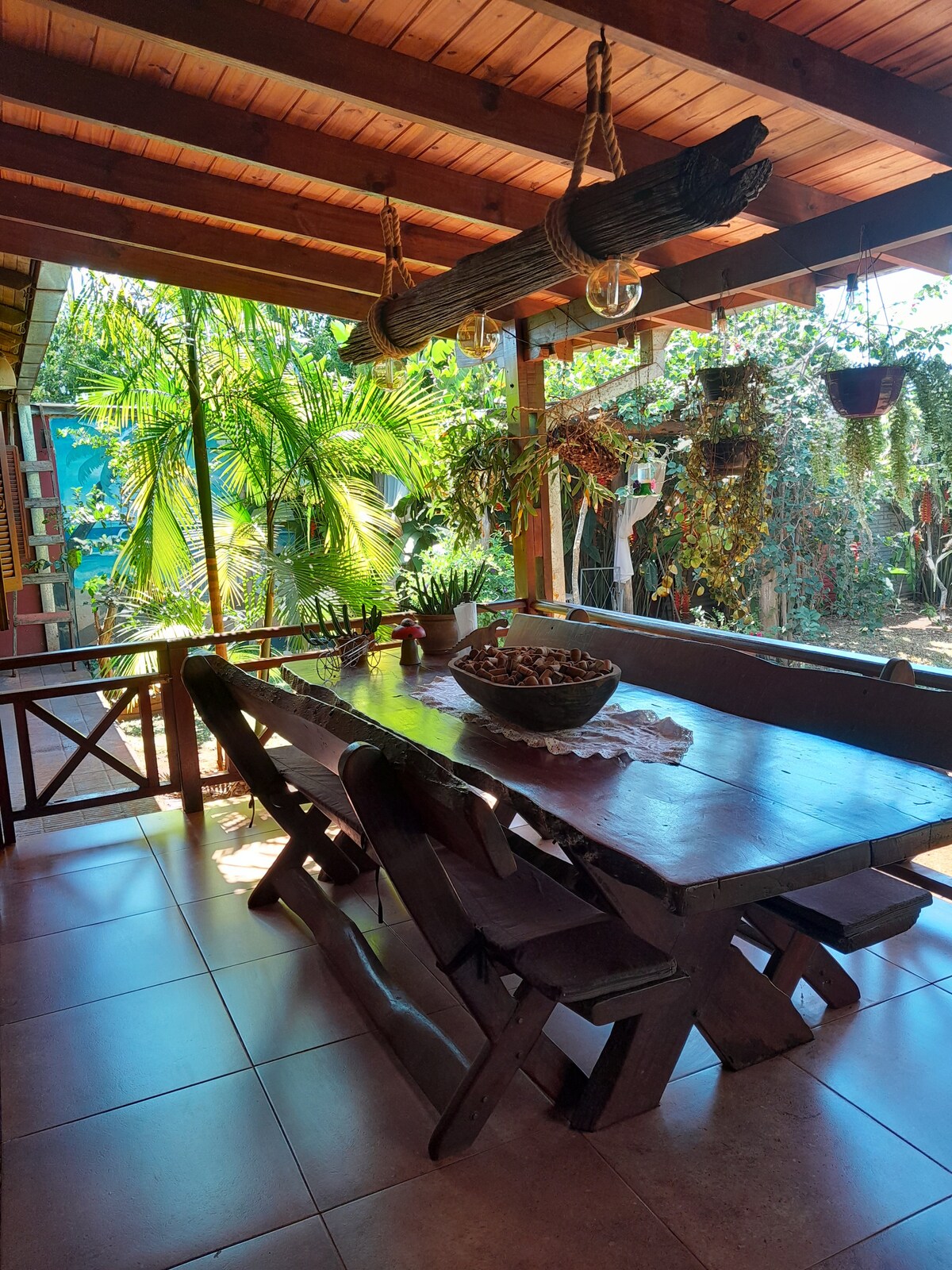 A spacious dining area features a large wooden table surrounded by six chairs. The natural light filters in through the open space, highlighting hanging plants and vibrant greenery that fills the yard. Beams of wood support the roof, creating a welcoming atmosphere.