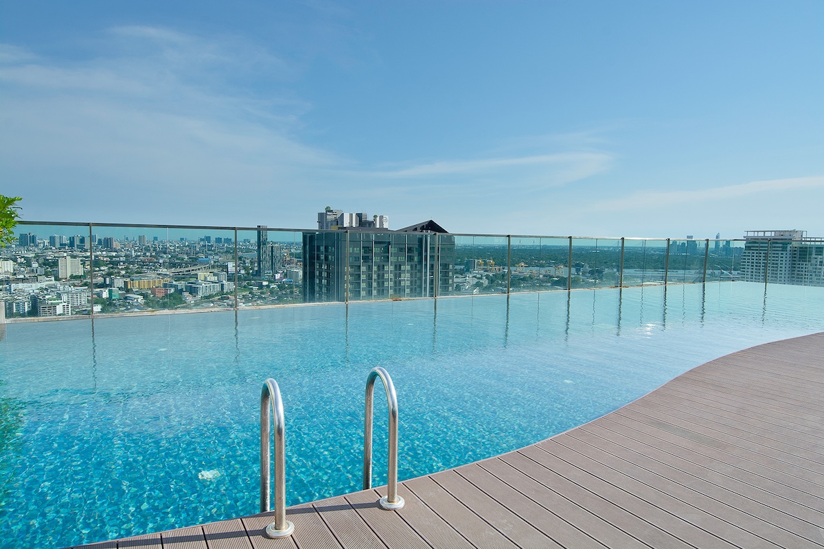 A rooftop pool is shown with clear blue water, reflecting the sky and surrounding skyline of Bangkok. Glass railings enhance the view, ensuring safety while enjoying the panoramic cityscape. Wooden decking surrounds the pool area, providing an inviting space for relaxation.
