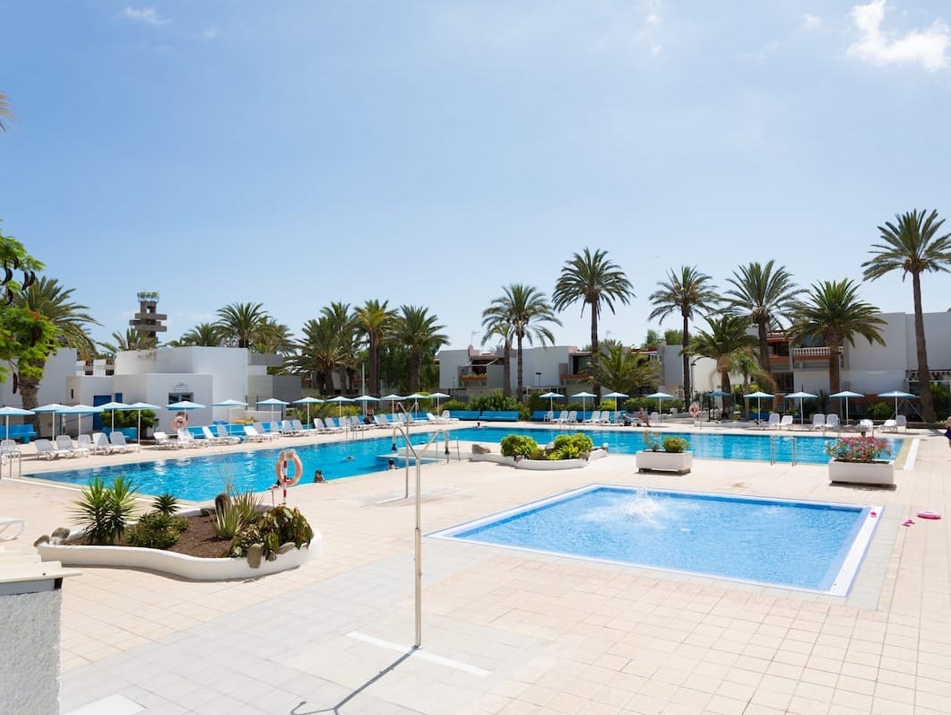 A serene communal pool area showcases two inviting swimming pools surrounded by well-maintained palm trees and sun loungers. A spacious deck area features vibrant landscaping, offering a relaxing atmosphere for guests to enjoy the sunny surroundings.