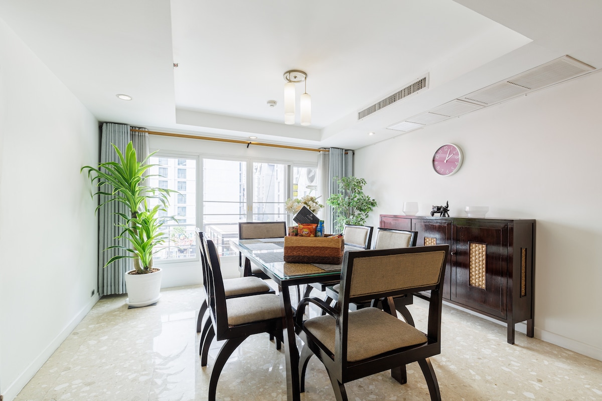 A spacious dining area is furnished with a large glass table surrounded by six wooden chairs. Natural light enters through large windows, complemented by indoor plants. A decorative bowl sits at the center of the table, enhancing the inviting decor.