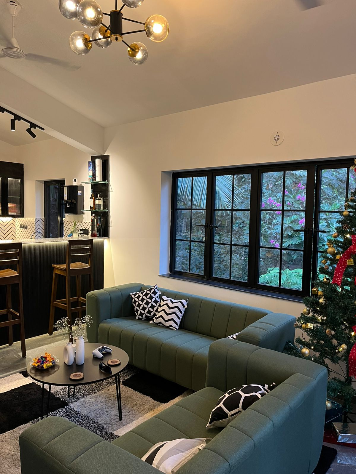 A living area features two green sofas arranged around a black coffee table, with decorative items placed on top. Large windows provide a view of greenery outside. In the background, a bar with seating and shelves is seen, adding to the vibrant ambiance.