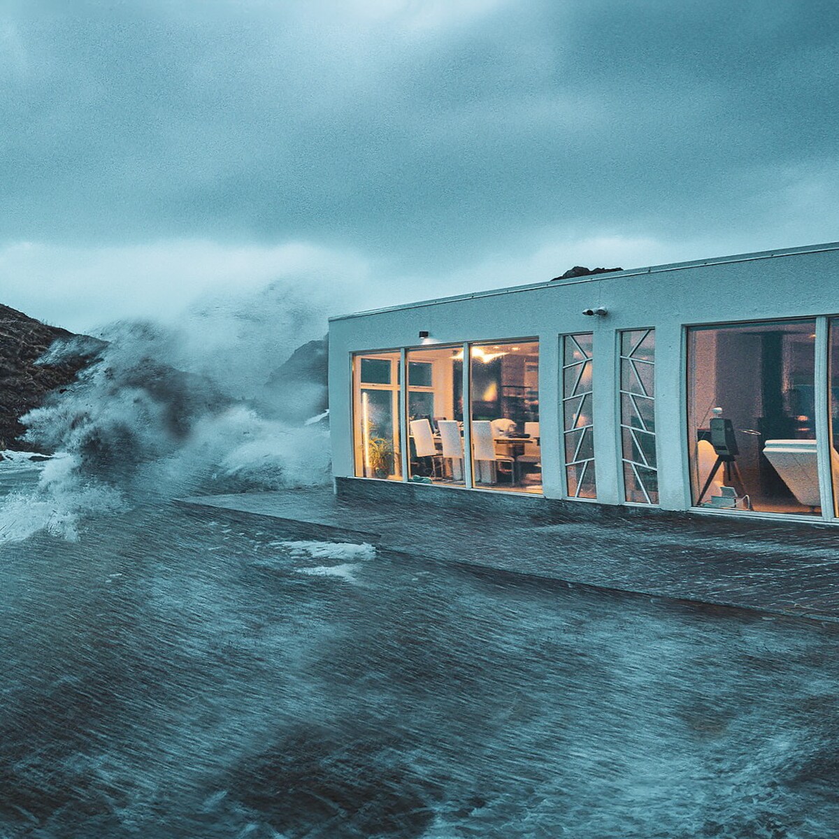 A modern villa is positioned close to a stormy sea, with large glass doors showcasing views of crashing waves. The exterior features sleek lines and a minimalist design, while soft lighting is visible inside, enhancing the inviting atmosphere despite the turbulent weather outside.