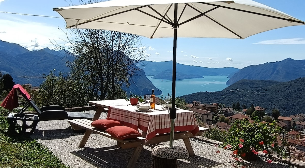 An outdoor dining area is set under a large umbrella, featuring a picnic table with a red and white checked tablecloth. Scenic views of distant mountains and a serene lake can be seen in the background. A comfortable lounge chair is also visible nearby.