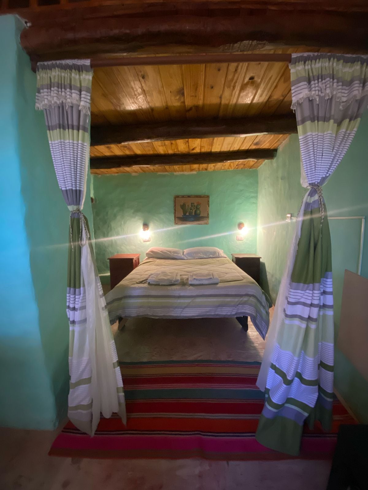 A cozy sleeping area features a spacious bed with soft linens. Vibrant green walls complement natural wood beams overhead. Light fixtures are positioned on either side of the bed, providing gentle illumination. A colorful woven rug enhances the warm ambiance of the space.