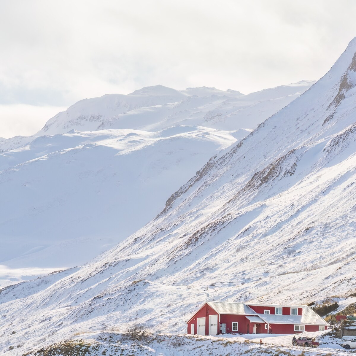 A red structure is situated on a snowy slope, surrounded by a mountainous landscape. The peaks are capped with snow and the sky is partially cloudy, creating a serene and tranquil winter scene.