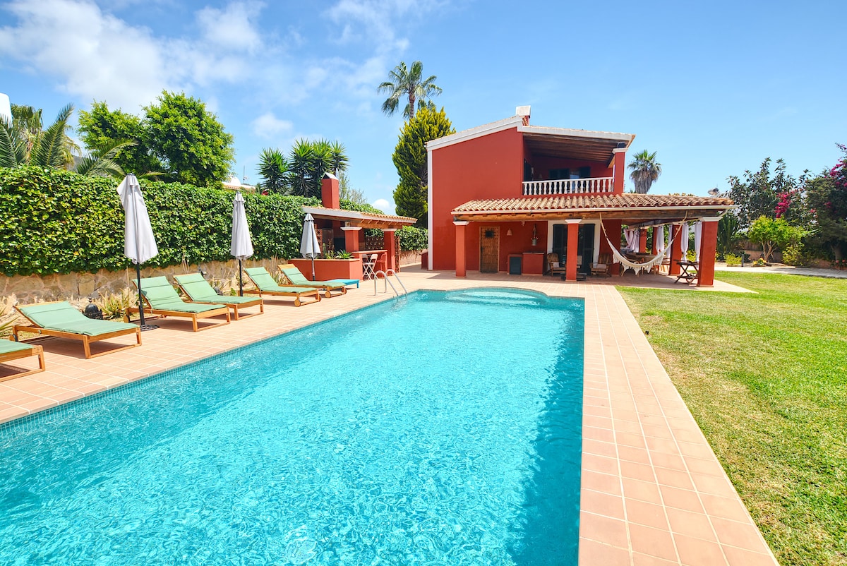A private swimming pool is surrounded by sun loungers under the shade of umbrellas. Lush greenery and palm trees frame the villa, which features a spacious outdoor dining area. The architecture includes a reddish exterior and a second-floor terrace, providing a tranquil environment.