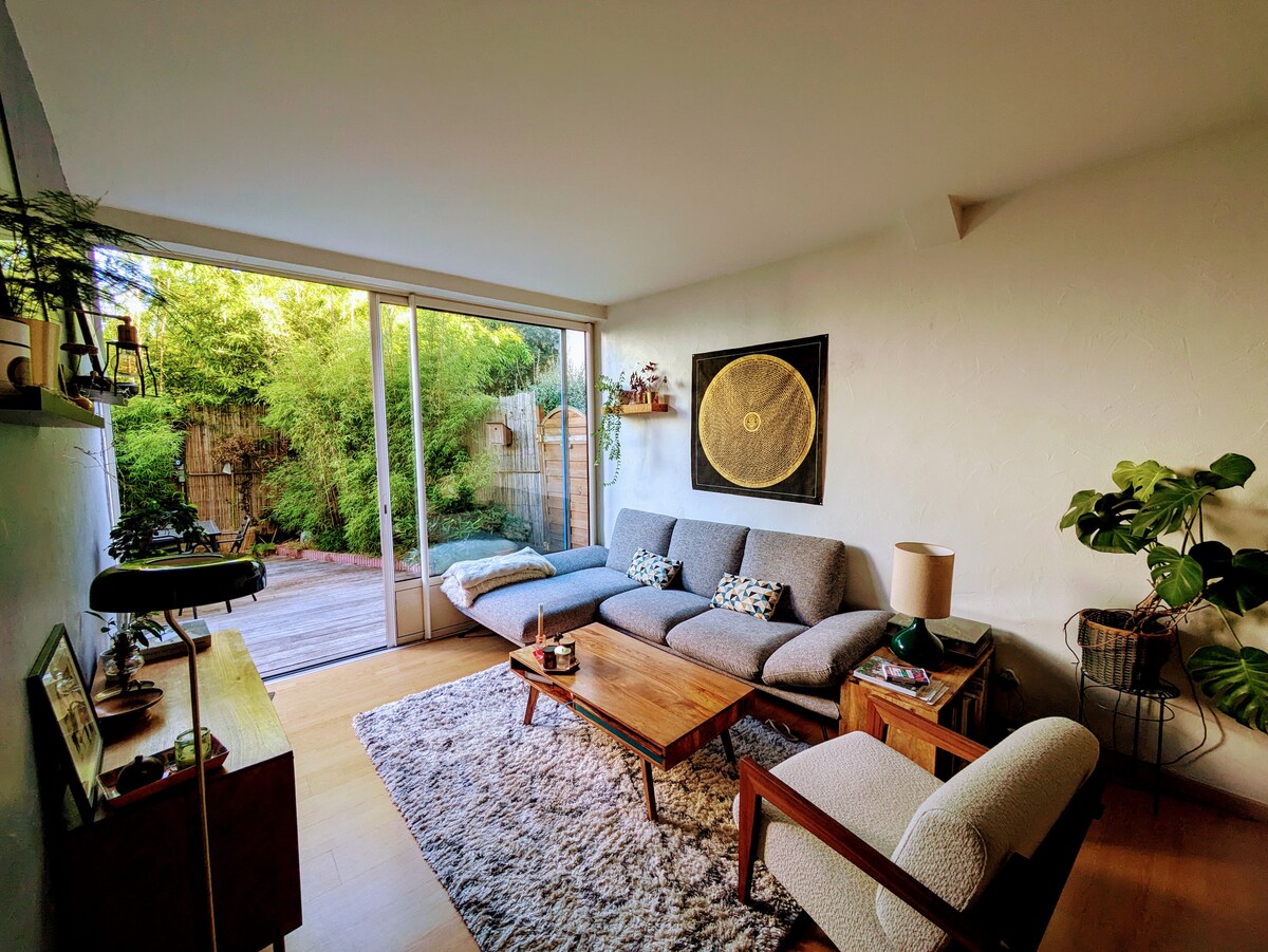 A bright living space includes a gray sofa and a wooden coffee table set on a soft area rug. Floor-to-ceiling glass doors open to a private garden area filled with greenery, enhancing the connection between indoor and outdoor environments.