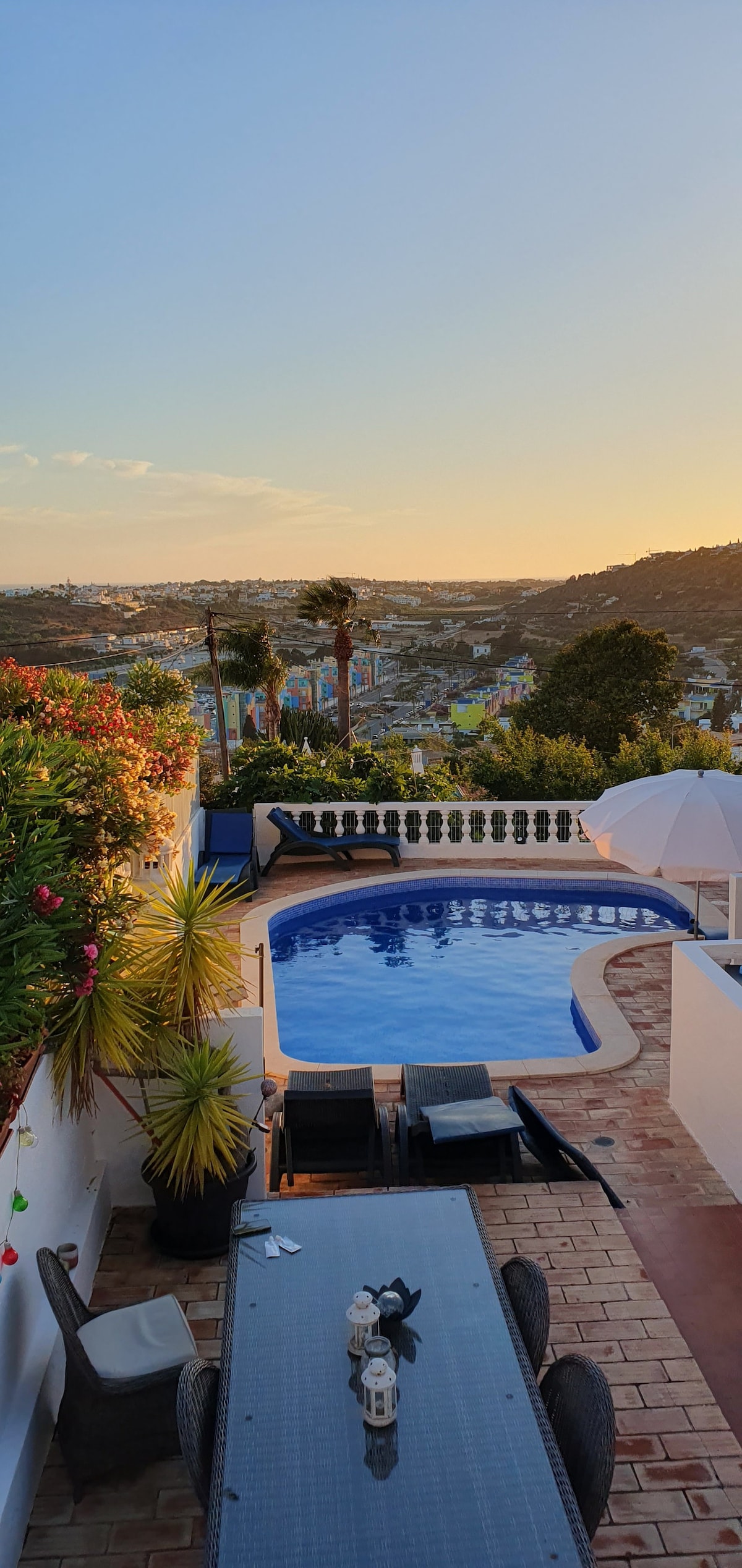 A view of the private pool surrounded by tropical plants, with a spacious outdoor dining table set for evening dining. The serene landscape displays the Albufeira Marina and mountains beyond, set against a colorful sunset sky.