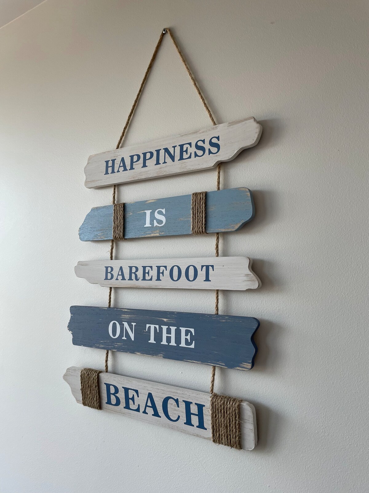 A decorative wall hanging is displayed, featuring wooden signs painted in white and blue tones. The signs contain the phrase 'Happiness is barefoot on the beach,' presented in a playful yet simple arrangement, adding a coastal charm to the space.