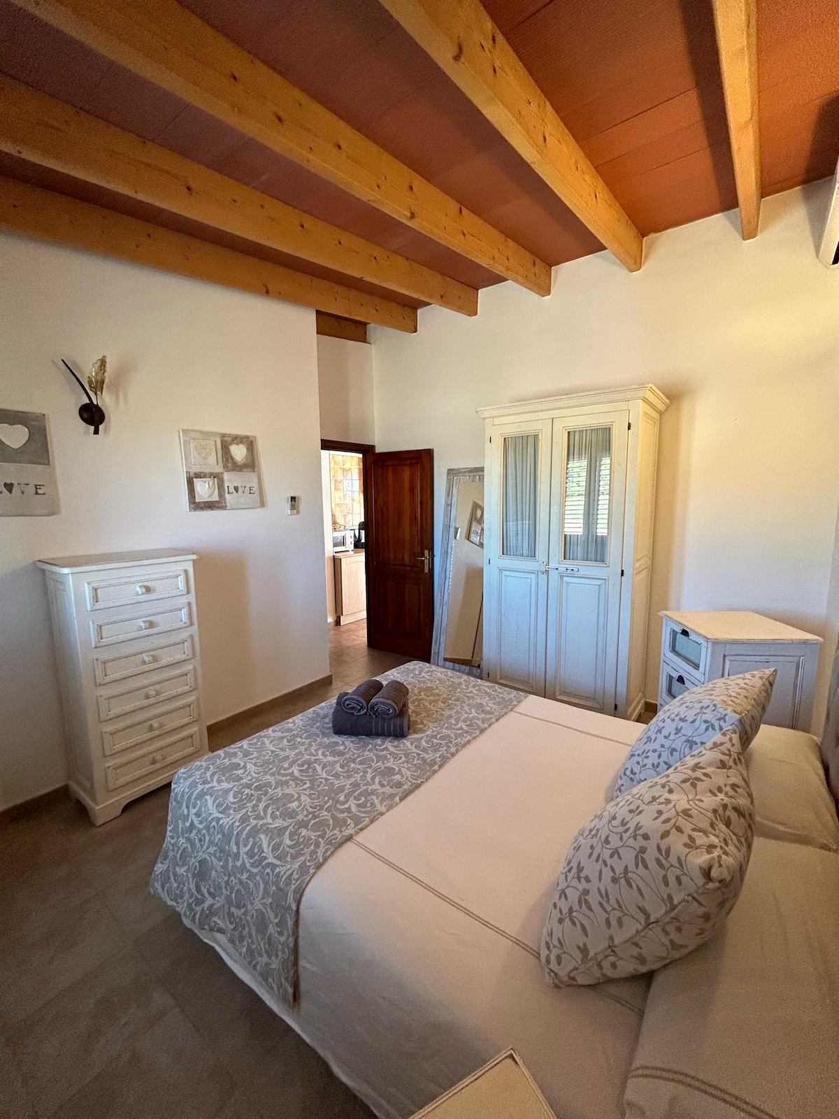 A cozy sleeping area is highlighted by wooden beams overhead. A light-colored bed is dressed with a patterned duvet and decorative pillows. Nearby, a white dresser and a cabinet provide storage, while natural light filters through a window with wooden shutters.