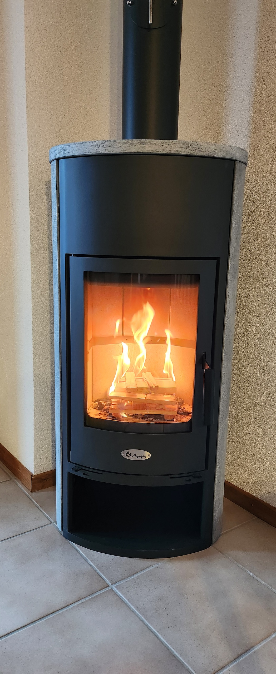 A modern wood stove is positioned against a neutral wall, featuring a tall, cylindrical design with a black and gray finish. Flames flicker warmly within the glass front, providing a comforting glow. The tiled flooring complements the stove's stylish appearance.