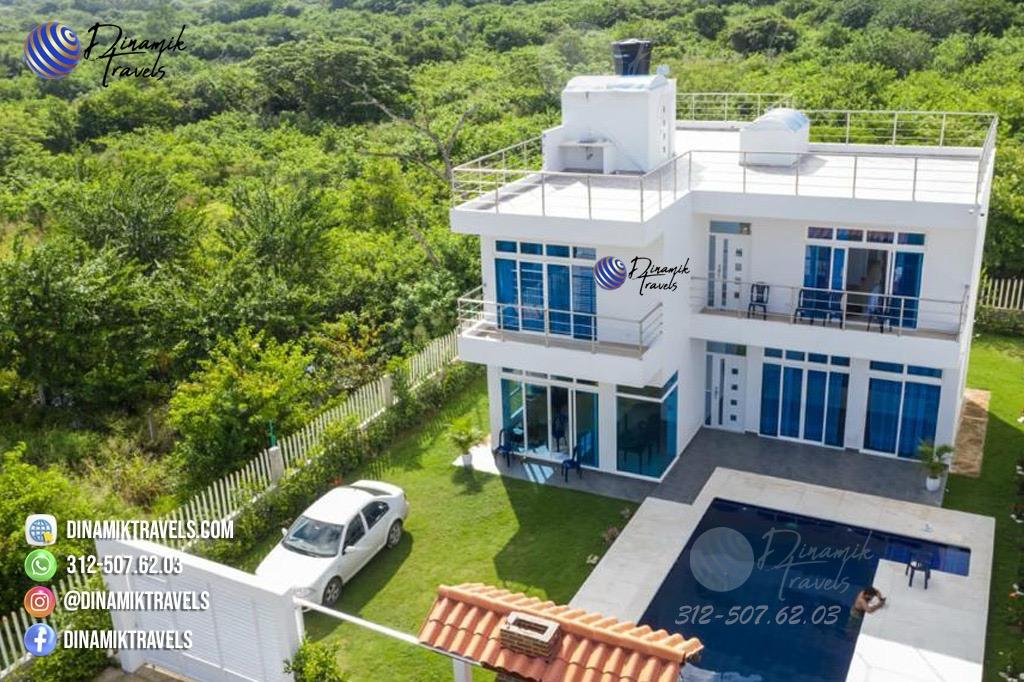 A modern two-story cabaña is presented with a private pool in the foreground. Lush greenery surrounds the property, offering a serene environment. Extensive windows invite natural light, while a rooftop terrace adds to the outdoor enjoyment. A vehicle is parked nearby, indicating convenient access.
