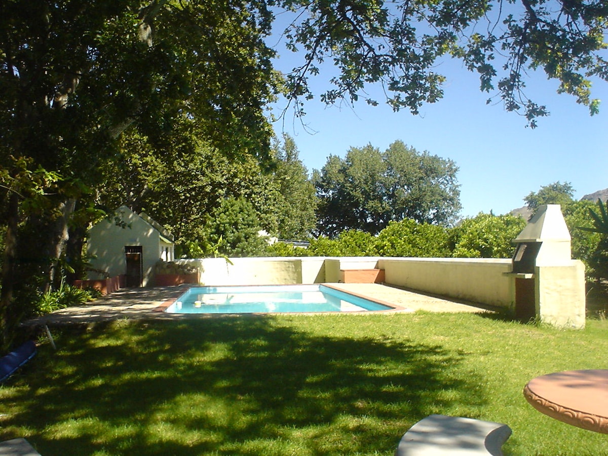 A swimming pool is set in a secluded area, surrounded by lush grass and shade from ancient oak trees. The pool area features a light-colored boundary wall, with a barbecue grill nearby. The backdrop includes well-maintained landscaping and clear blue skies.