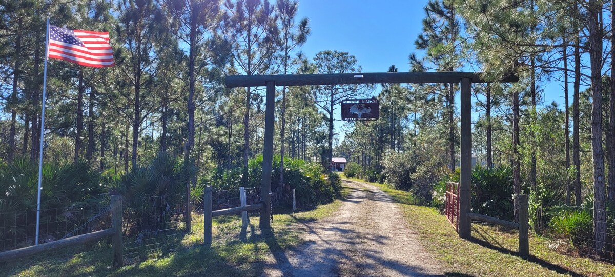 An entrance gate marks the beginning of the property, framed by trees and shrubs. A clear gravel path leads toward the farmhouse, with a sign visible above the gate. A flag waves gently to the side, adding a welcoming touch to the scene.
