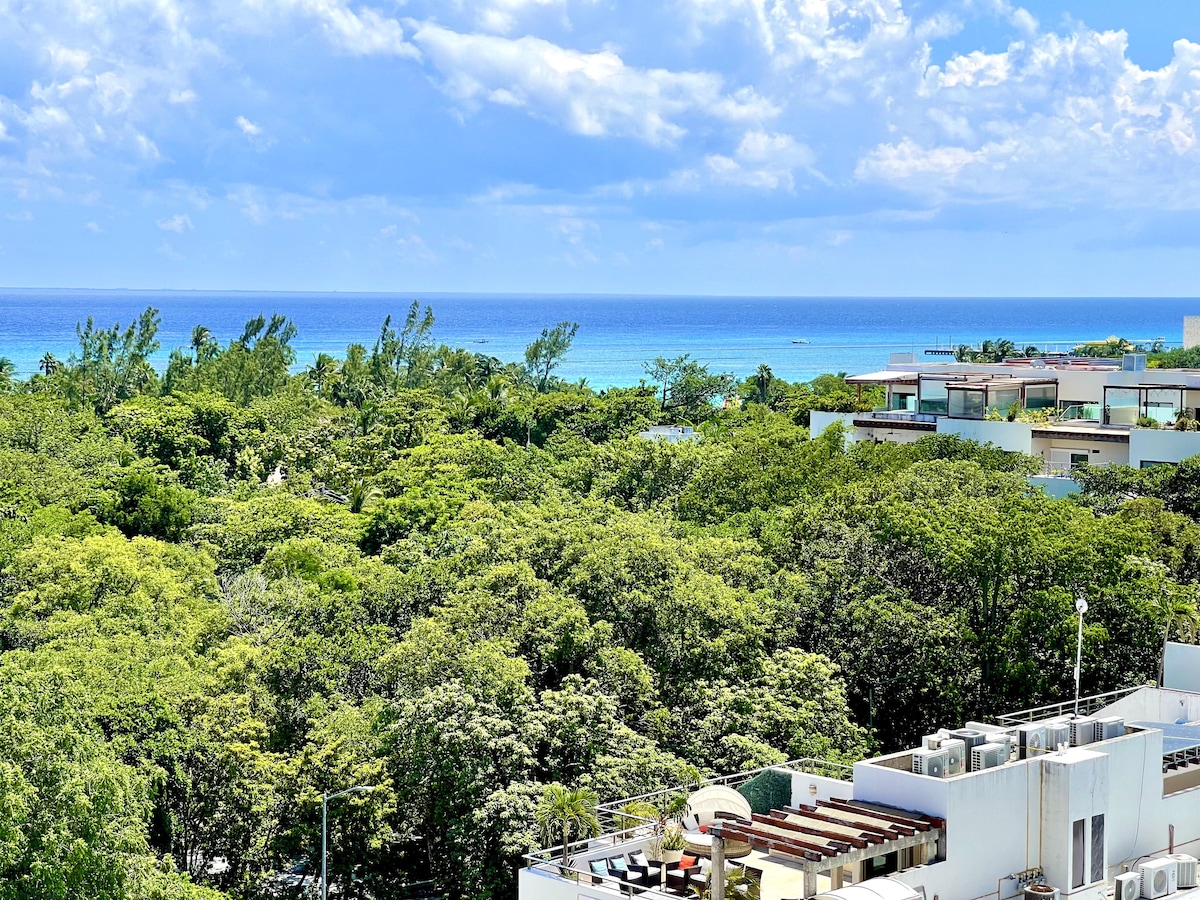 A scenic view captures the lush greenery surrounding the property, with glimpses of the turquoise Caribbean Sea visible in the distance. Fluffy clouds drift across the bright blue sky, enhancing the overall tranquil atmosphere of the environment.