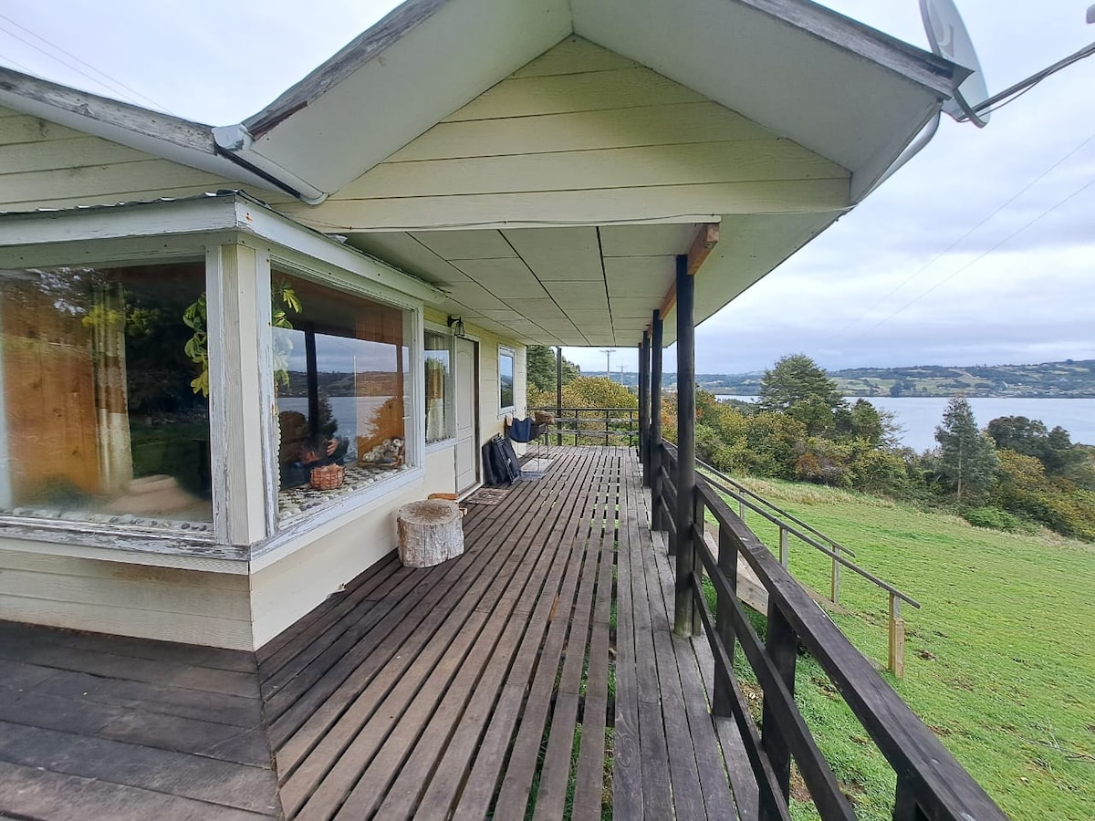 A spacious deck extends from the cabin, featuring wooden flooring and railings. Comfortable seating arrangements provide views of the surrounding natural landscape and the water. Large windows allow natural light to enter the interior, enhancing the connection with the outdoors.