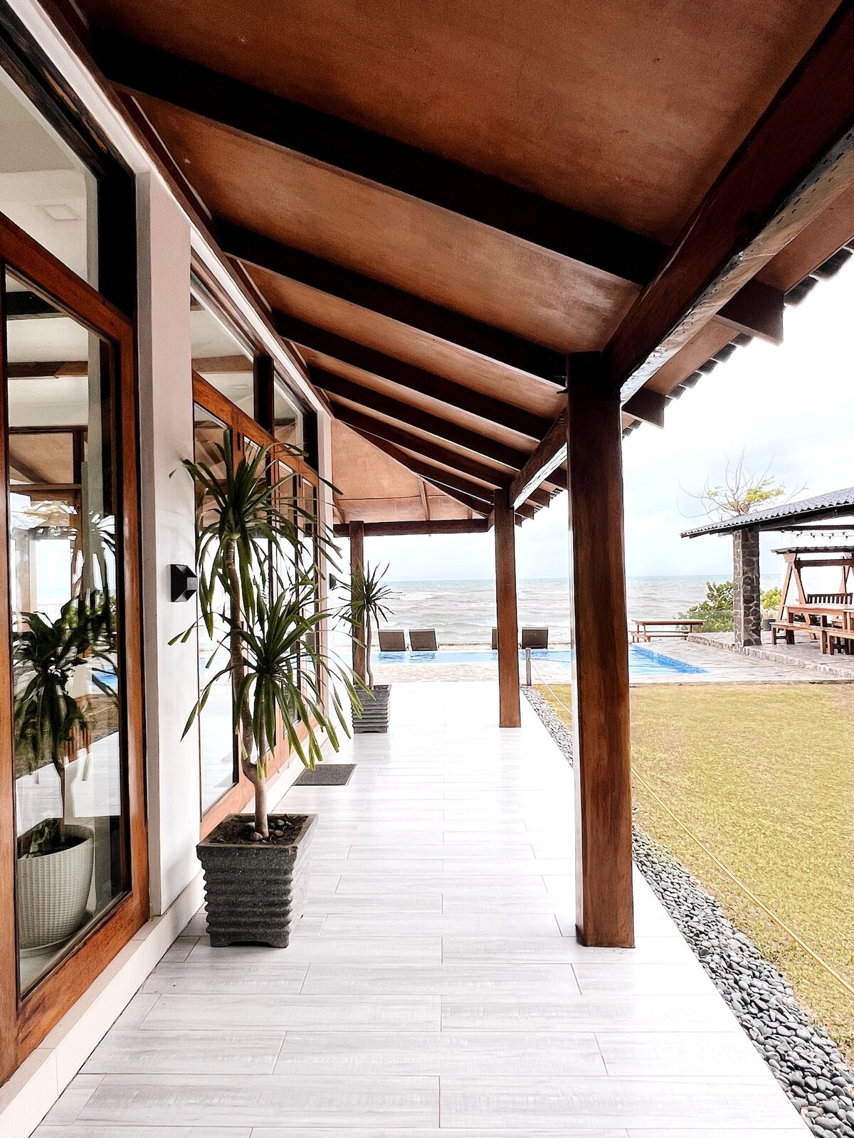 A veranda features wooden beams and a tiled floor. Potted plants are arranged along the walls, providing a natural touch. Large glass windows allow views of the beach and pool area, enhancing the connection with the outdoor environment.