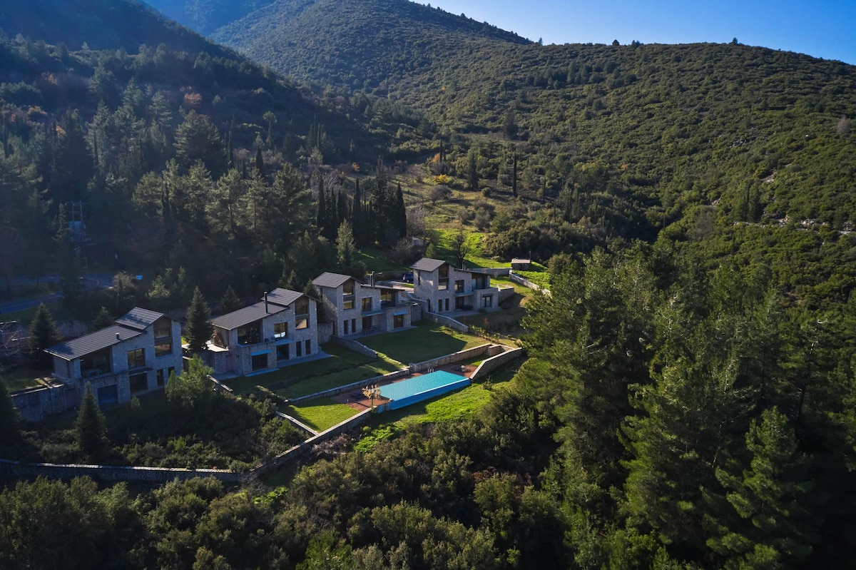 An aerial view showcases Alti Luxury Villa nestled in a lush green landscape. The expansive property includes a shared outdoor swimming pool and well-manicured gardens, all surrounded by rolling hills and trees, offering a seamless blend of nature and luxury.