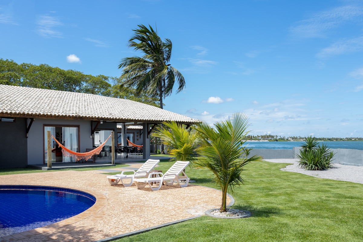 A serene outdoor area features a heated pool surrounded by well-maintained grass. Lounge chairs are positioned for relaxation, while palm trees gently sway in the breeze. The house, with large glass doors, overlooks the water, providing a peaceful coastal setting.