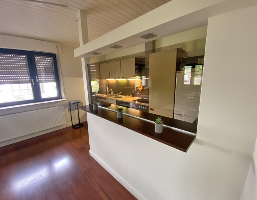 The open kitchen features a sleek design with modern cabinetry and appliances. A wooden countertop separates it from the living area, adorned with small potted plants. Natural light filters through the dual windows, highlighting the warm wood flooring.
