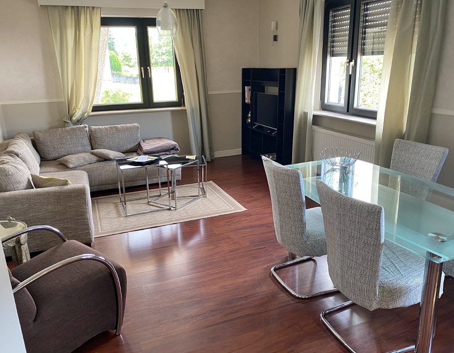 A living area with a light brown sofa and accent chairs is paired with a glass dining table surrounded by grey upholstered chairs. Natural light floods through two windows, which are framed by soft, light curtains, providing a comfortable and inviting space.