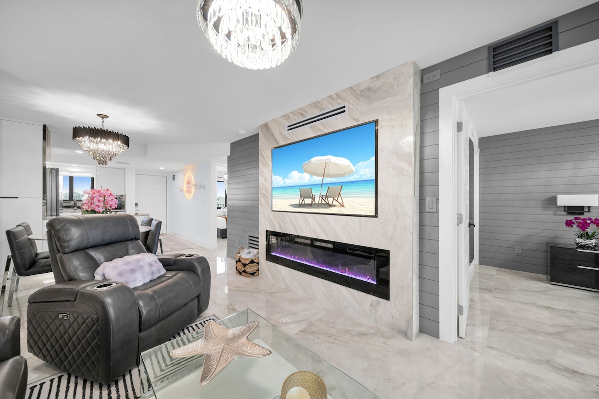 The living area features a sleek leather sofa and recliners, complemented by a modern coffee table. A wall-mounted 70" 4K Smart HDTV is framed by a contemporary fireplace insert. Soft lighting from a decorative chandelier enhances the space, which is finished with elegant marble flooring.