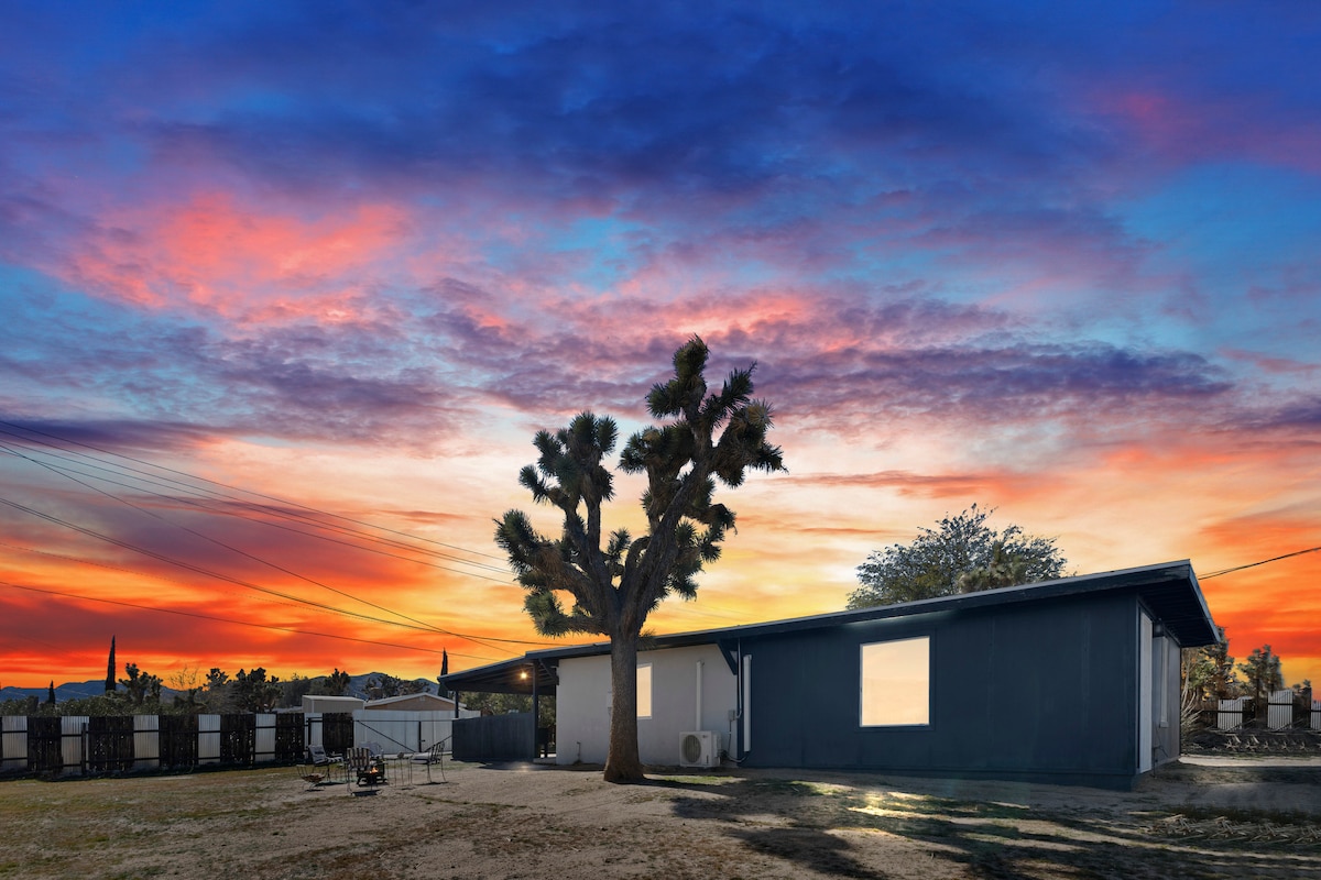 A spacious back unit is viewed against a vibrant sunset sky, featuring rich hues of orange, purple, and blue. A single large Joshua tree stands in the foreground, highlighting the natural desert landscape.