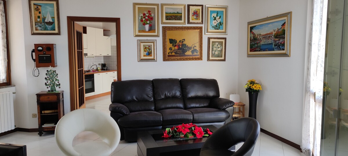 The living area is furnished with a dark leather sofa and two modern chairs surrounding a low table adorned with decorative flowers. The walls feature an assortment of framed artwork, and natural light streams in through a window draped with sheer curtains.