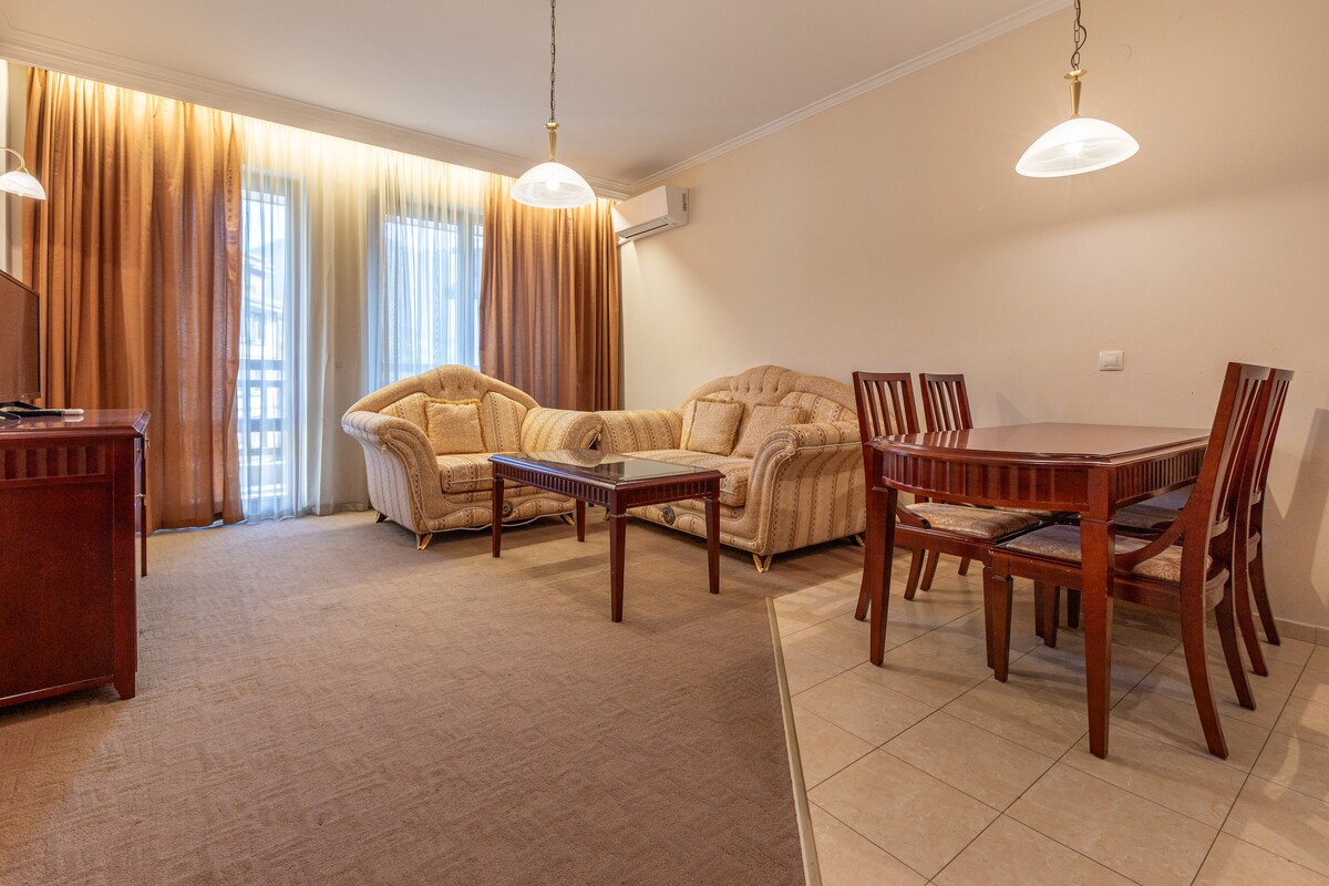 A spacious living area features two beige couches and a wooden coffee table. Natural light enters through large glass doors, illuminating the room. A dining table with six wooden chairs is positioned beside the tiled floor, creating a comfortable and inviting environment.