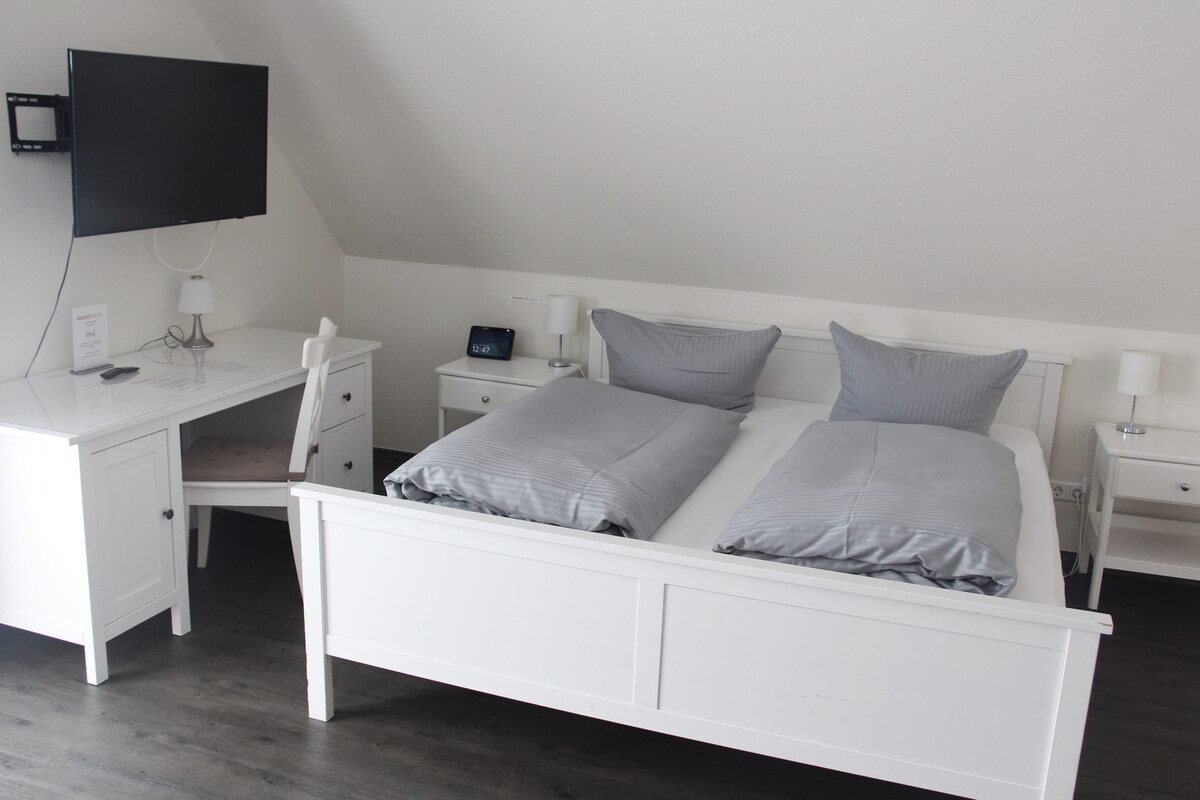 A modern studio space features a spacious bed with two comforters, complemented by bedside tables with lamps. A large TV is mounted on the wall opposite the bed. A desk with a chair is situated beside a drawer unit, providing additional storage and workspace.