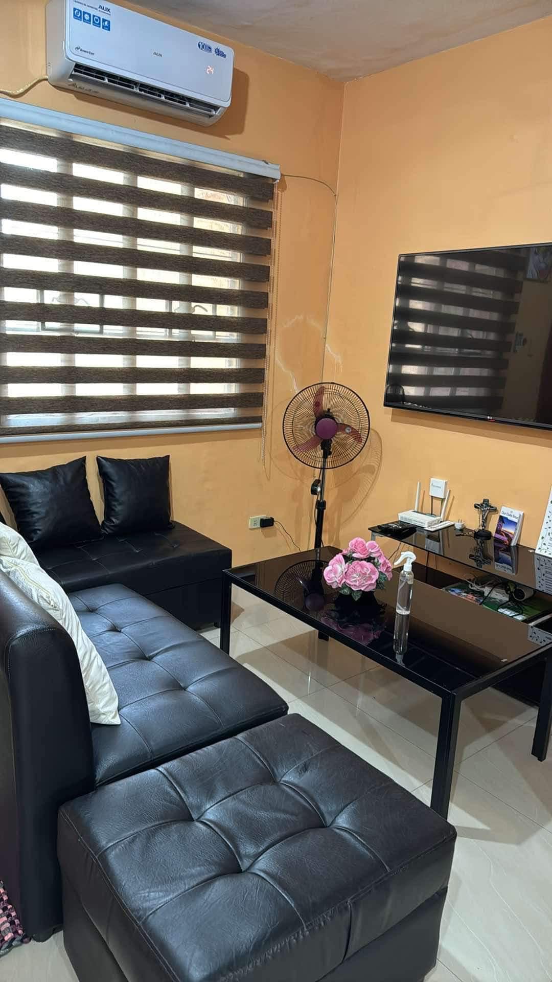 A cozy living area features a black sectional sofa with decorative cushions and a sleek black coffee table. An air-conditioning unit is mounted above, while a wall-mounted TV is positioned across from the seating. Natural light filters through the horizontal blinds, creating a warm ambiance.