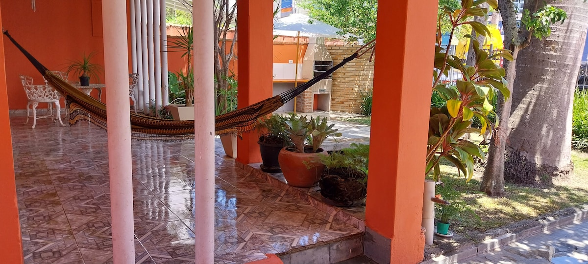 A covered patio area is highlighted by a relaxed hammock suspended between two sturdy columns. Potted plants surround the space, adding greenery. The tiled floor reflects natural light, and a spacious outdoor area can be seen in the background, offering room for leisure activities.