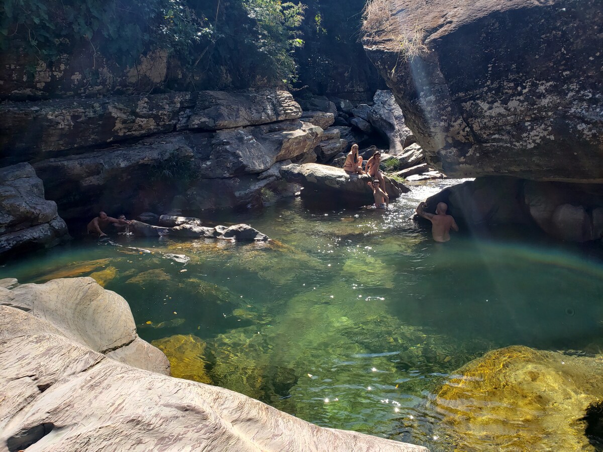 A natural swimming area is surrounded by large, rugged boulders, with clear green water reflecting sunlight. Several people are seen enjoying the serene environment, some seated on rocks near the water. Lush vegetation is present, contributing to the tranquil and rustic feel of the landscape.