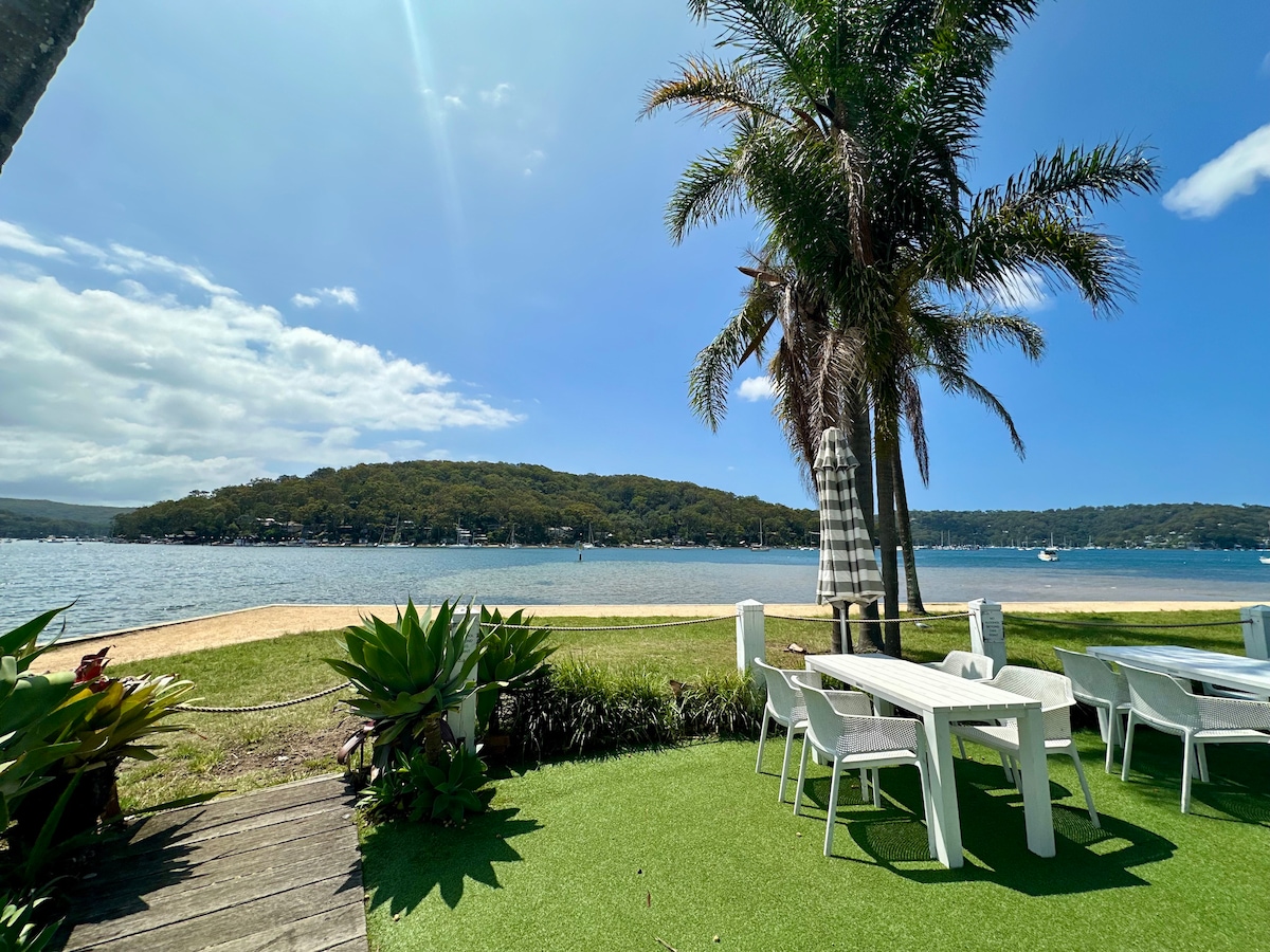A scenic view of Pittwater is showcased, featuring sandy shores and gentle waves. Lush greenery and palm trees frame the image, complemented by a dining set placed on well-maintained grass. The backdrop includes hills covered in trees, contributing to the serene atmosphere.