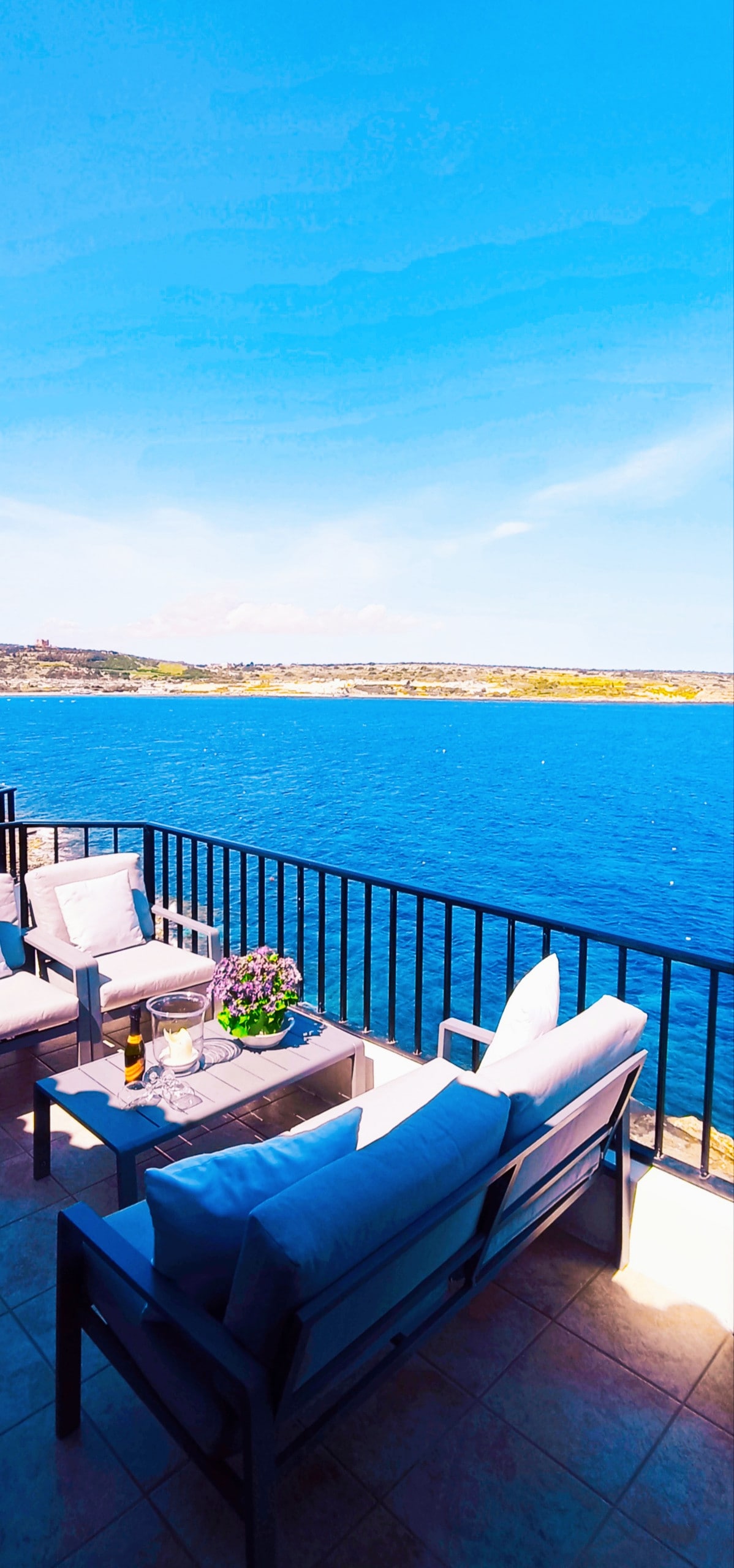 A balcony view presents a spacious seating area with comfortable chairs and a coffee table overlooking a vibrant blue sea. The horizon features lush land, enhancing the tranquil atmosphere. A small potted plant adds a touch of greenery to the outdoor space, bathed in natural light.