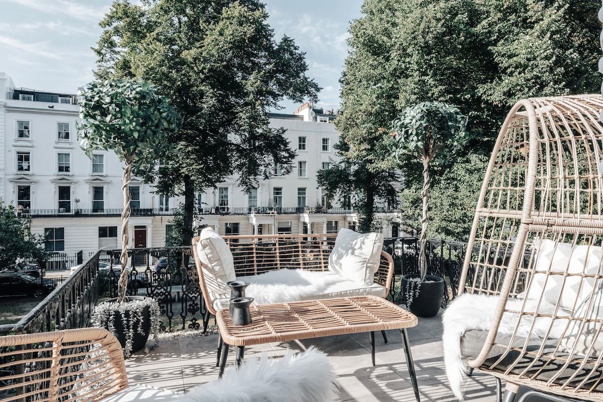 A serene outdoor terrace features a comfortable seating area, with woven furniture and soft cushions. Lush greenery surrounds the space, providing a tranquil atmosphere. Views of the historic buildings and garden square can be seen in the background, enhancing the inviting outdoor environment.