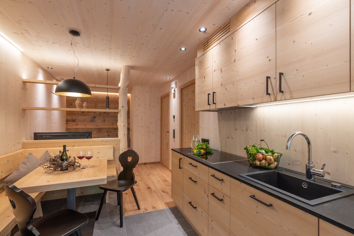 A wooden kitchen area is highlighted by natural wood cabinetry and a dark countertop. A cozy dining table with surrounding chairs is positioned beside a wooden bench. Soft lighting enhances the ambiance, while a bowl of fresh produce sits prominently on the counter.