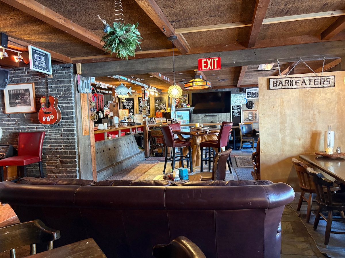 The spacious common area features a mix of seating options, including leather couches and wooden tables. A well-lit bar area is visible in the background along with a large television. Decorative elements, such as guitars and signs, create a relaxed atmosphere for guests.