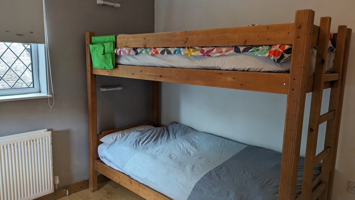 A spacious bedroom features two standard wooden bunk beds. Each bed is made with simple bedding, while a window allows natural light to fill the room. A radiator can be seen beneath the window, and the walls are painted in neutral tones.