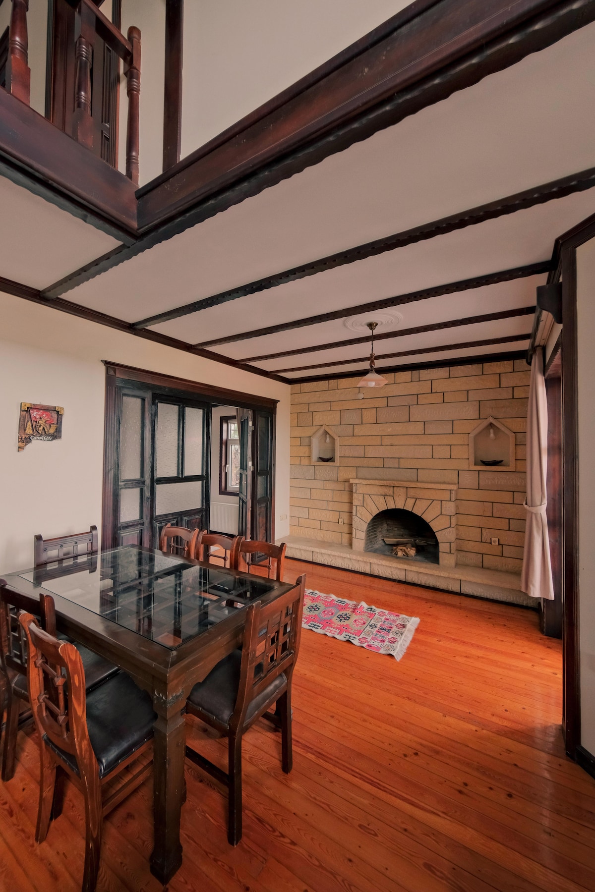 A dining area is presented with a wooden table surrounded by six chairs. A large window allows natural light to fill the space, and a traditional stone fireplace serves as a focal point against the wall. The ceiling features dark wooden beams.