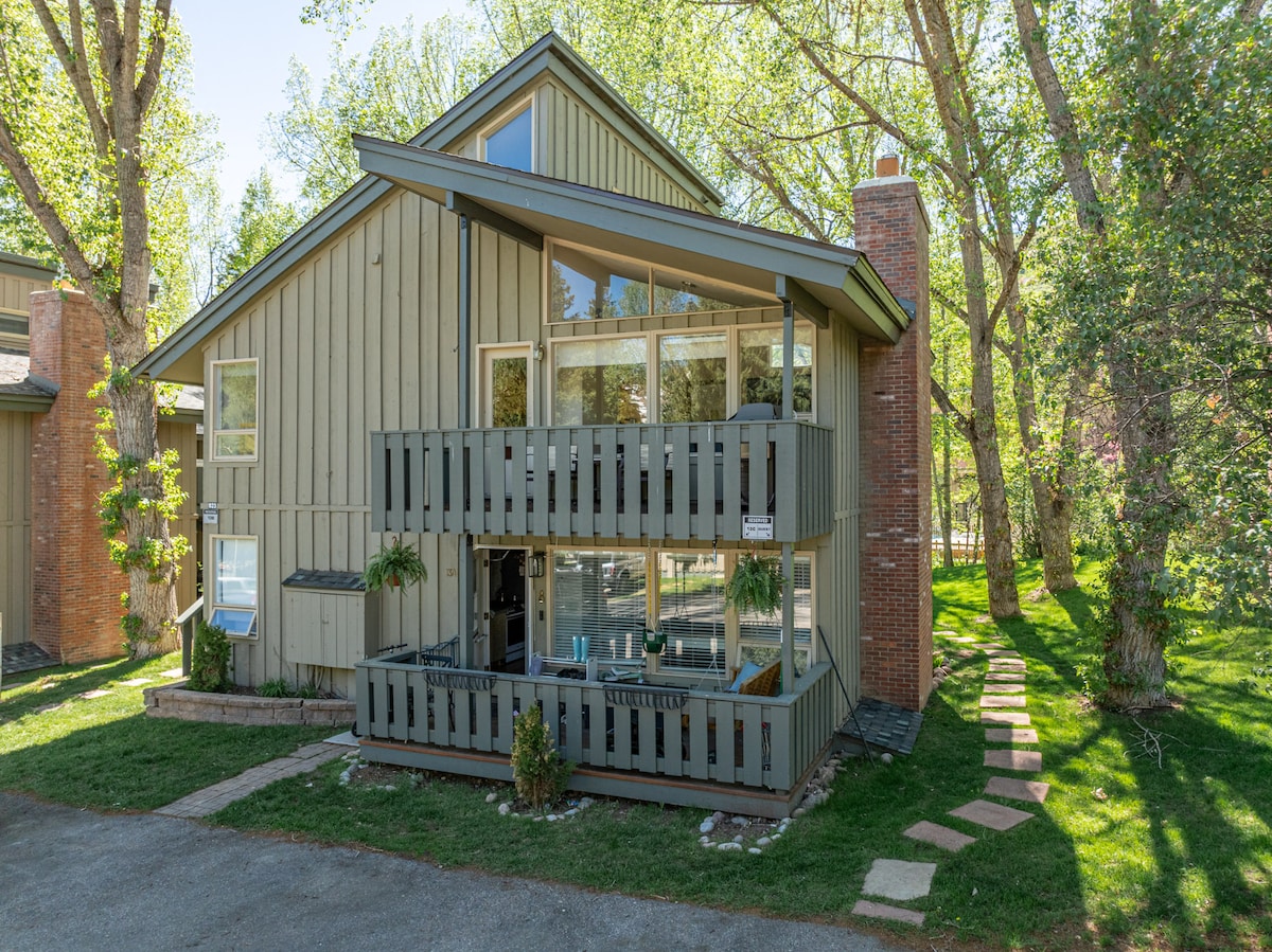 The exterior of the condominium showcases a two-story structure with gray siding and large windows. A wraparound balcony features seating areas and is surrounded by lush greenery. Stone pathways lead to the entrance, enhancing the inviting outdoor space near the trees.