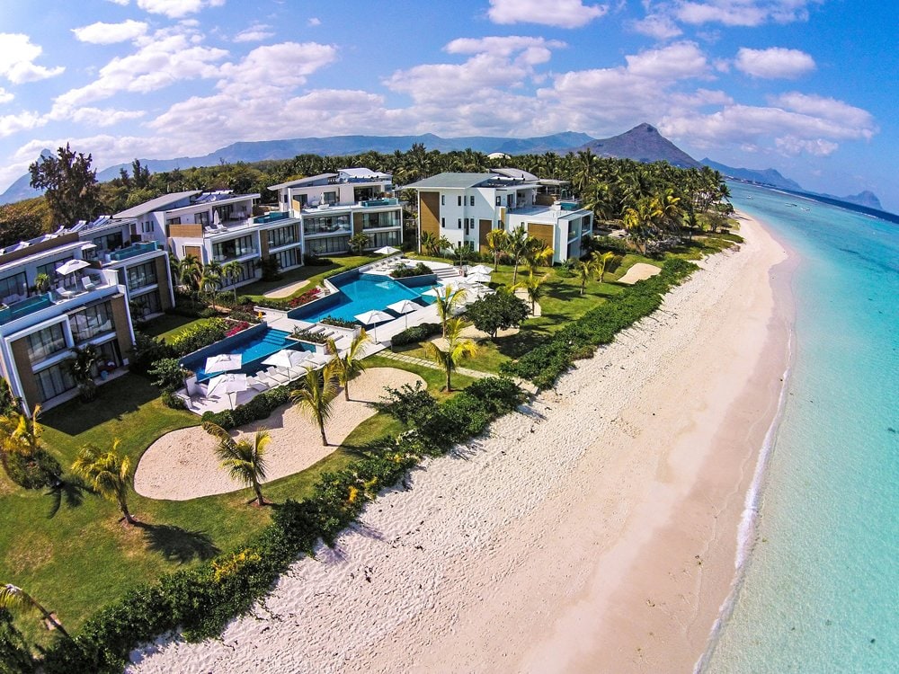 The beachfront property features a landscaped area with lush palm trees and a clear view of the sandy beach. Swimming pools are visible within the complex, surrounded by spacious lawns. The azure ocean extends towards the horizon, enhancing the serene coastal environment.