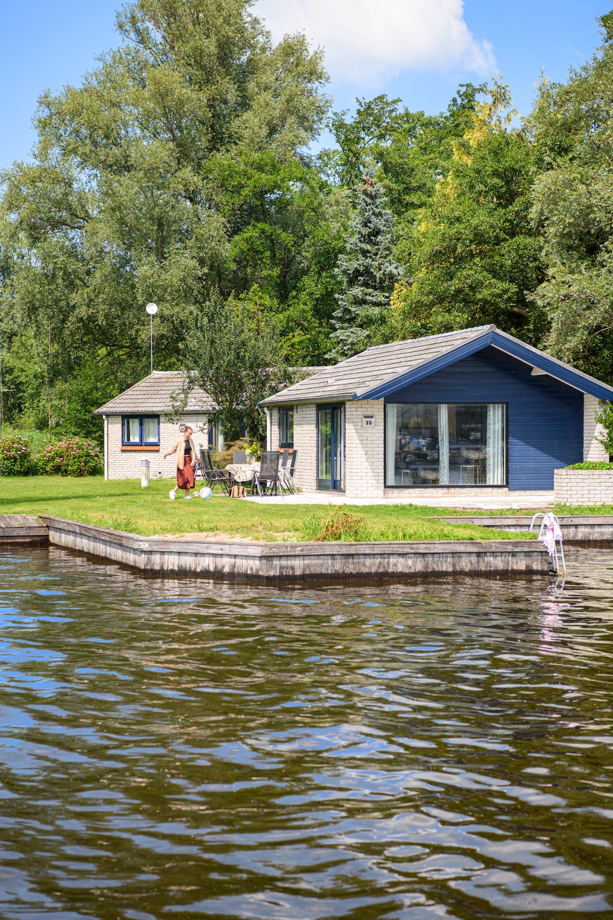 A bungalow is positioned on the banks of the water, featuring large glass doors that open to a grassy area. Outside furniture is arranged on the terrace, with trees providing shade in the background. The serene water reflects the surrounding greenery.