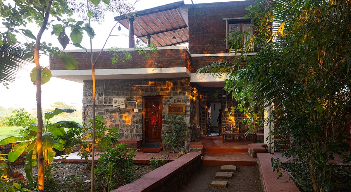 The main entrance of the property features a blend of stone and brick architecture, surrounded by lush greenery. A pathway leads to the door, lined with decorative steps and plants. Natural light filters through the trees, creating dappled patterns on the exterior.