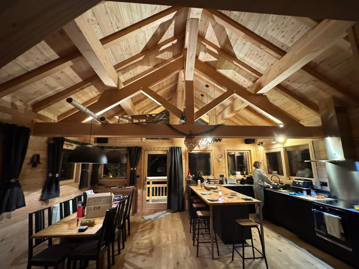 A spacious kitchen area is highlighted by wooden beams and large windows, providing ample natural light. A central kitchen island is surrounded by several high-backed stools. Black cabinetry and appliances complement the light wood tones, while contrasting dark curtains frame the windows.